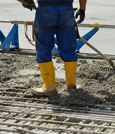 Eine Person mit blauer Hose und gelben Gummistiefeln steht auf einer Baustelle im frischen Beton. Im Vordergrund sind Stahlarmierungen zu sehen, im Hintergrund eine Maschine zur Betonverarbeitung.

                        